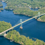 000 Islands Bridge spanning over the St. Lawrence River, surrounded by lush green trees under a partly cloudy sky, on a serene semi-sunny day.