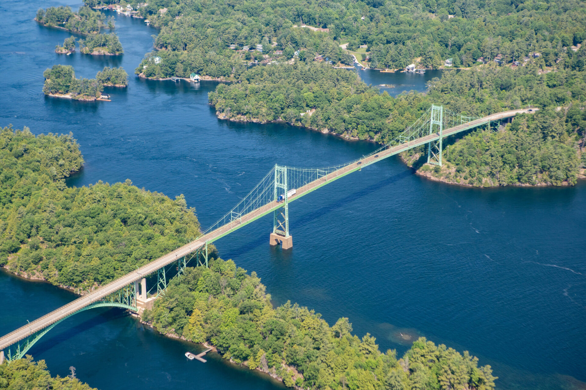 000 Islands Bridge spanning over the St. Lawrence River, surrounded by lush green trees under a partly cloudy sky, on a serene semi-sunny day.