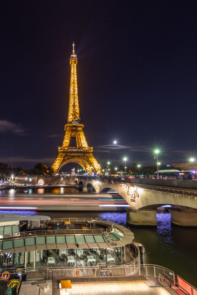 Long exposure photograph of the Eiffel Tower at night, with a boat's light trail visible on the Seine River.