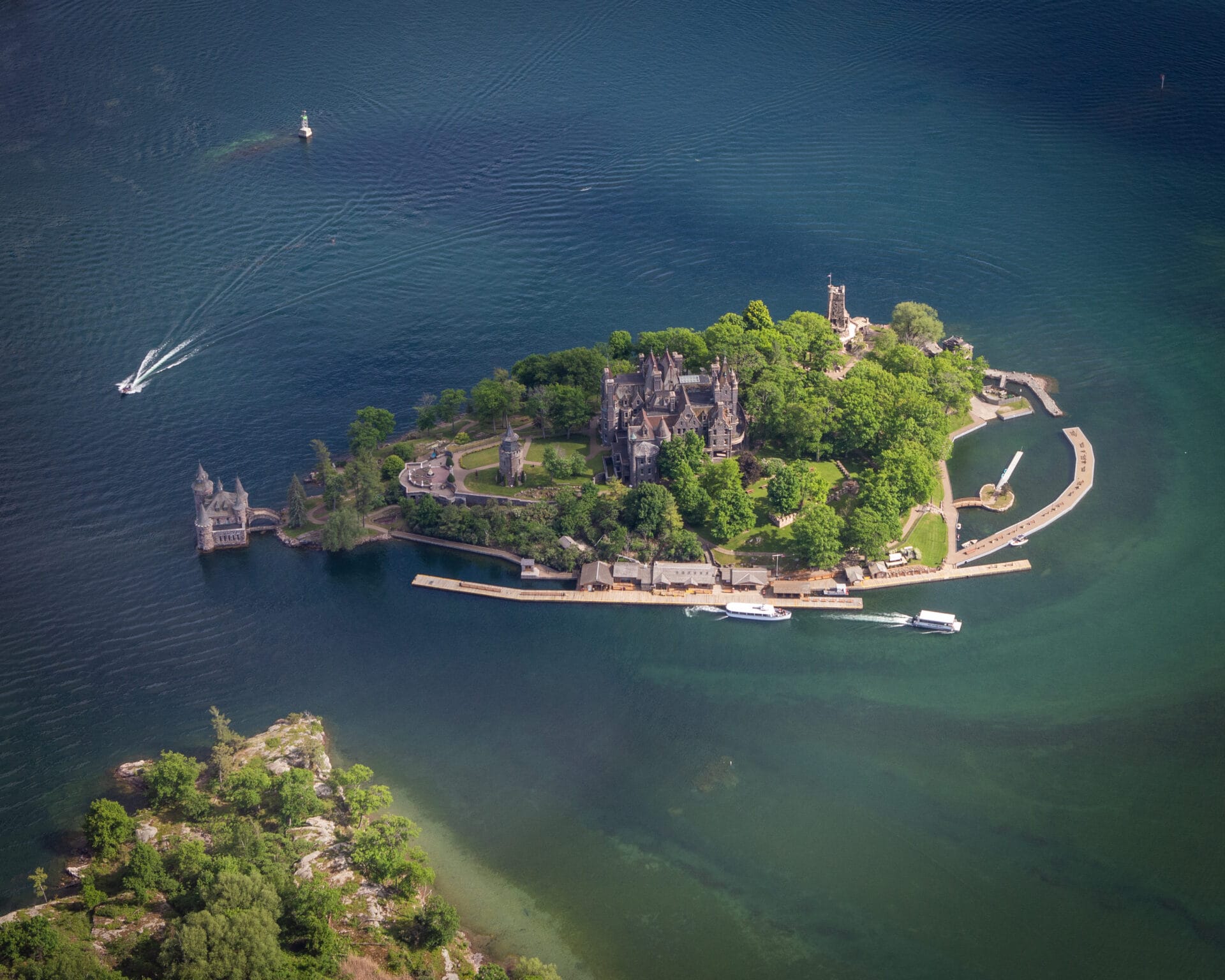 A picturesque view of Boldt Castle, an impressive historic mansion on Heart Island in the Thousand Islands region. The castle features elegant stone architecture, multiple turrets, and lush gardens surrounded by the sparkling waters of the St. Lawrence River.